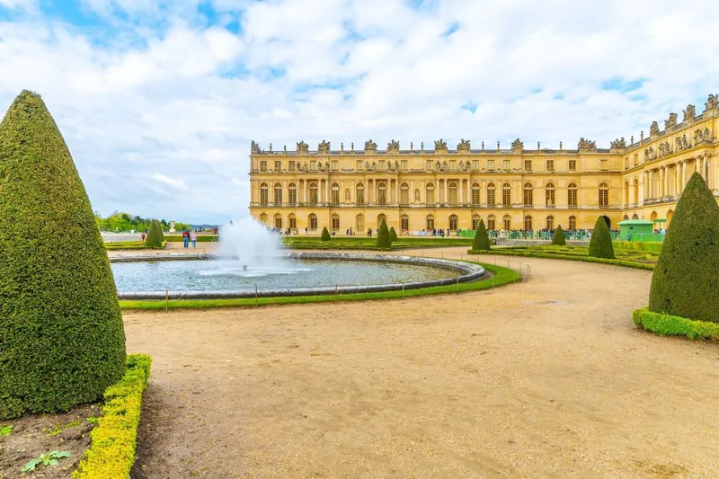Round fountain pool with topiary cones and the garden facade of Versailles palace