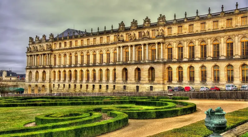 The garden front of the Palace of Versailles above clipped box parterres, with cars parked beside the wing